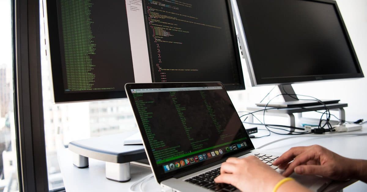 A developer typing on a laptop in front of code-filled monitors.